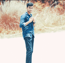 a young man wearing a blue plaid shirt and black pants stands in front of tall grass