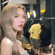 a woman with blonde hair is standing in front of a mirror in a room with a man in a yellow shirt behind her .