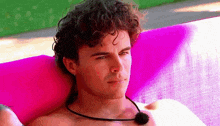 a shirtless man with curly hair is laying on a pink blanket with a microphone around his neck .