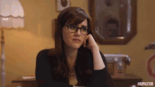 a woman wearing glasses is sitting at a table with her hand on her chin and looking at the camera .