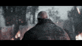 a bald man in a black coat stands in front of a burning building