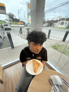 a young man sitting at a table holding a plate of food and wearing a shirt that says colinas