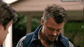 a man with a beard and a vest is standing in front of a house .