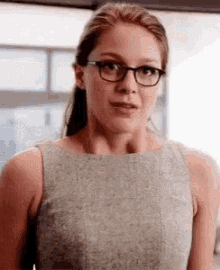 a woman wearing glasses and a grey dress is standing in front of a window and looking at the camera .