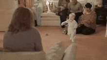 a baby is walking in a living room with a group of people watching