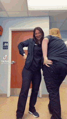 two women in scrubs are standing next to each other in front of a bathroom sign .