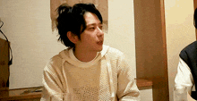 a man wearing a white sweater and a black vest is sitting in a room