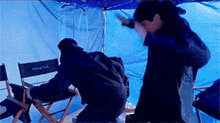 a man in a director 's chair is standing next to another man in front of a blue tarp .