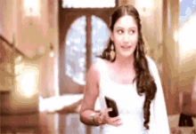 a woman in a white dress is standing in a room holding a cell phone in her hand .
