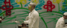 a man with yellow paint on his face is playing drums in front of a graffiti covered wall .