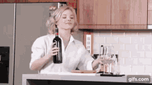 a woman in a white shirt is holding a bottle of wine and a glass of wine in a kitchen .