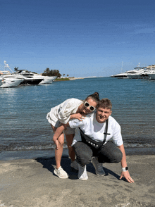 a man and a woman are posing for a picture on the beach with boats in the background