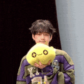 a young man in a purple sweater is holding a yellow stuffed animal with a face on it .
