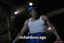 a man in a white tank top and blue shorts says richardovo ego in a dark room