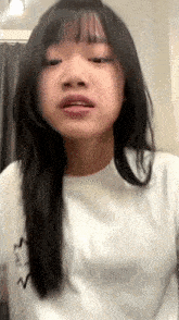 a girl with long black hair is wearing a white t-shirt and making a funny face .