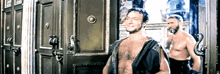 two shirtless men are standing in front of a door and one has the letter e on his shoulder