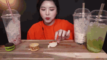 a woman in an orange shirt is eating a sandwich next to two cups of drinks