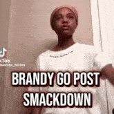 a young man wearing a hat and a white shirt is standing in front of a door and says brandy go post smackdown