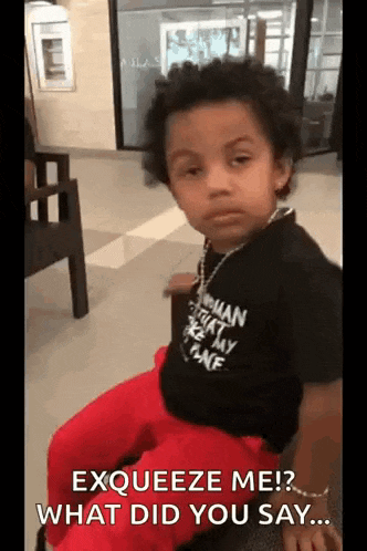 a young boy wearing a black shirt and red pants is sitting on a chair and making a funny face .