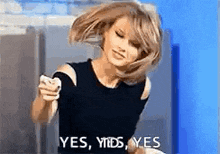 a woman in a black top is holding a cell phone and says yes , yes , yes .