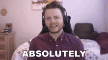 a man wearing headphones and a purple shirt is sitting in front of a bed and saying `` absolutely '' .