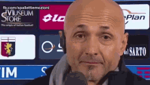a bald man is talking into a microphone in front of a museum store banner