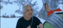 two men are standing in the snow talking to each other . one of the men is wearing a helmet .