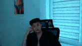 a young man sitting in front of a window with a painting of a woman behind him