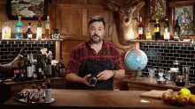 a man in a plaid shirt holds a shaker in front of a globe and bottles of liquor