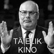 a man with glasses and a beard holds his hands up in front of a sign that says taielik kino