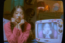 a girl in a pink sweater is sitting in front of a tv that has a picture of her on it