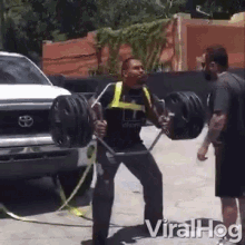 a man is lifting a barbell in front of a truck while another man watches .
