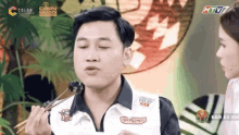 a man is eating food with chopsticks on a television show while a woman looks on .
