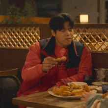 a man in a red jacket is sitting at a table eating chicken
