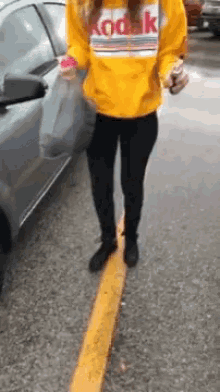 a woman in a yellow kodak sweatshirt is standing on the side of the road next to a car .