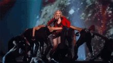a woman in a red jacket is standing on a stage surrounded by a group of dancers .