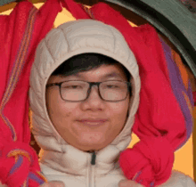 a young man wearing glasses and a hooded jacket holds a red scarf around his neck