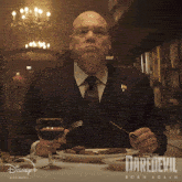 a man is sitting at a table with a plate of food and a glass of wine in front of a poster for daredevil