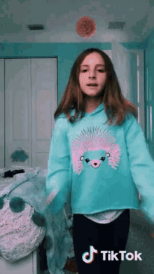 a girl wearing a blue sweater with a hedgehog on it stands in front of a bed with a tiktok sticker