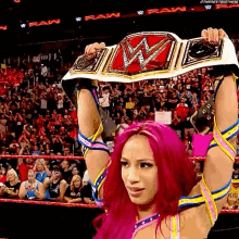 a woman with pink hair is holding a wrestling championship belt over her head in a ring .