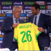 a man in a suit holds up a yellow shirt that says ancelotti 26