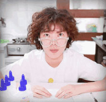 a woman wearing glasses sits at a table with a bottle of glue