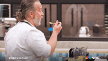 a man with a ponytail is eating something from a spoon with the nbc logo behind him