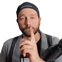 a man with a beard wearing a under armour hat holds his finger to his mouth .