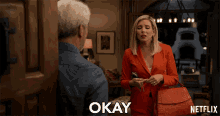 a woman in a red suit is standing next to a man and the word okay is on the screen behind her