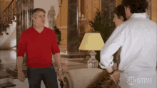 a man in a red sweater is standing in a living room talking to a woman and a man in a white shirt .