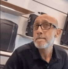 a bald man with glasses and a beard is sitting in front of a shelf with luggage .