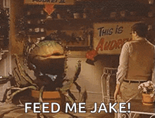 a man is standing in front of a carnivorous plant that says feed me jake .