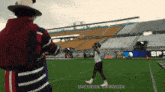 a man in a hat stands on a football field with the word shedeur sanders on the bottom right