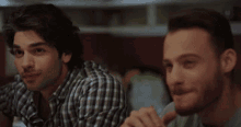 two men are sitting at a table talking to each other . one of the men is wearing a plaid shirt .